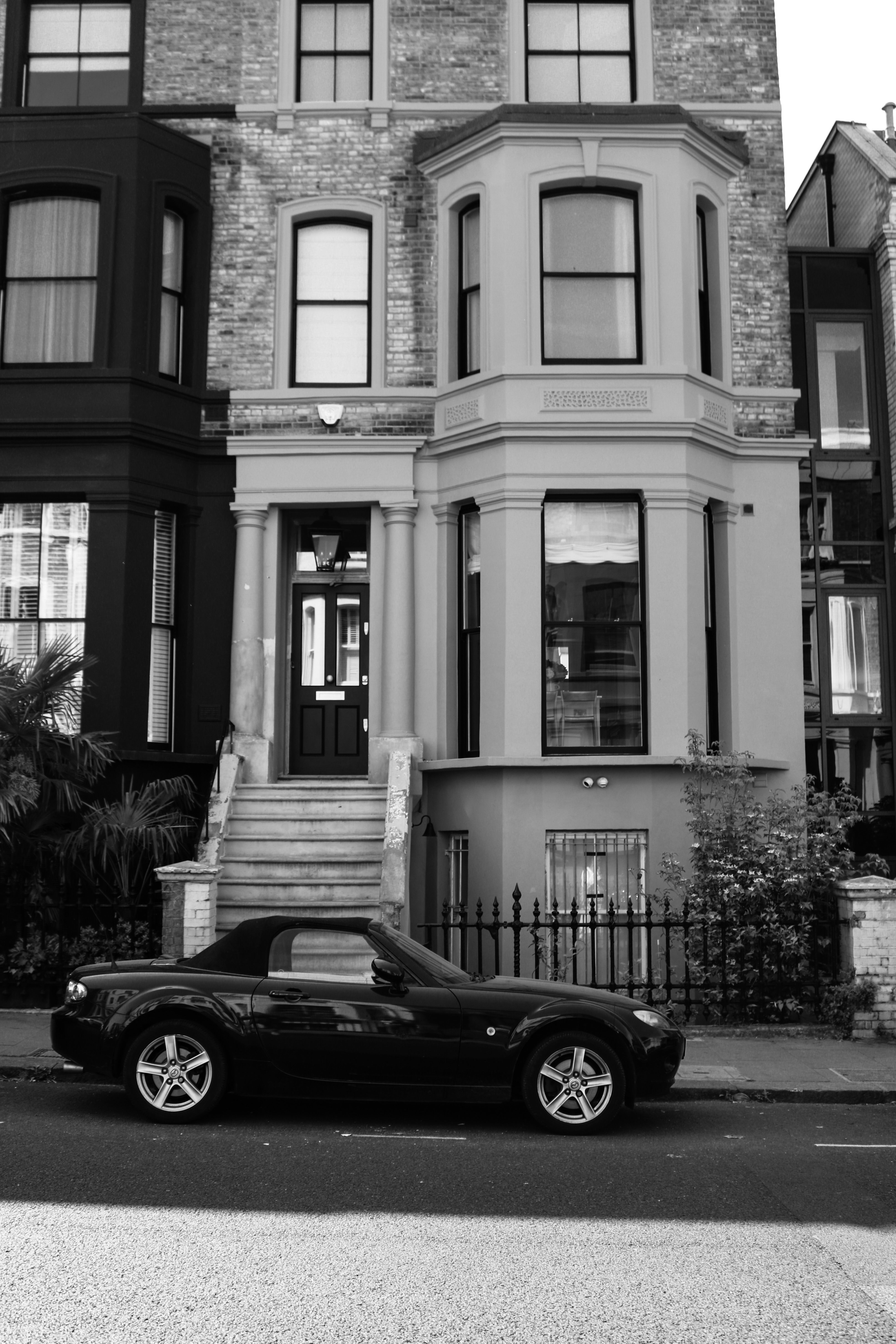 A sleek black convertible car parked outside a classic urban townhouse in black and white.