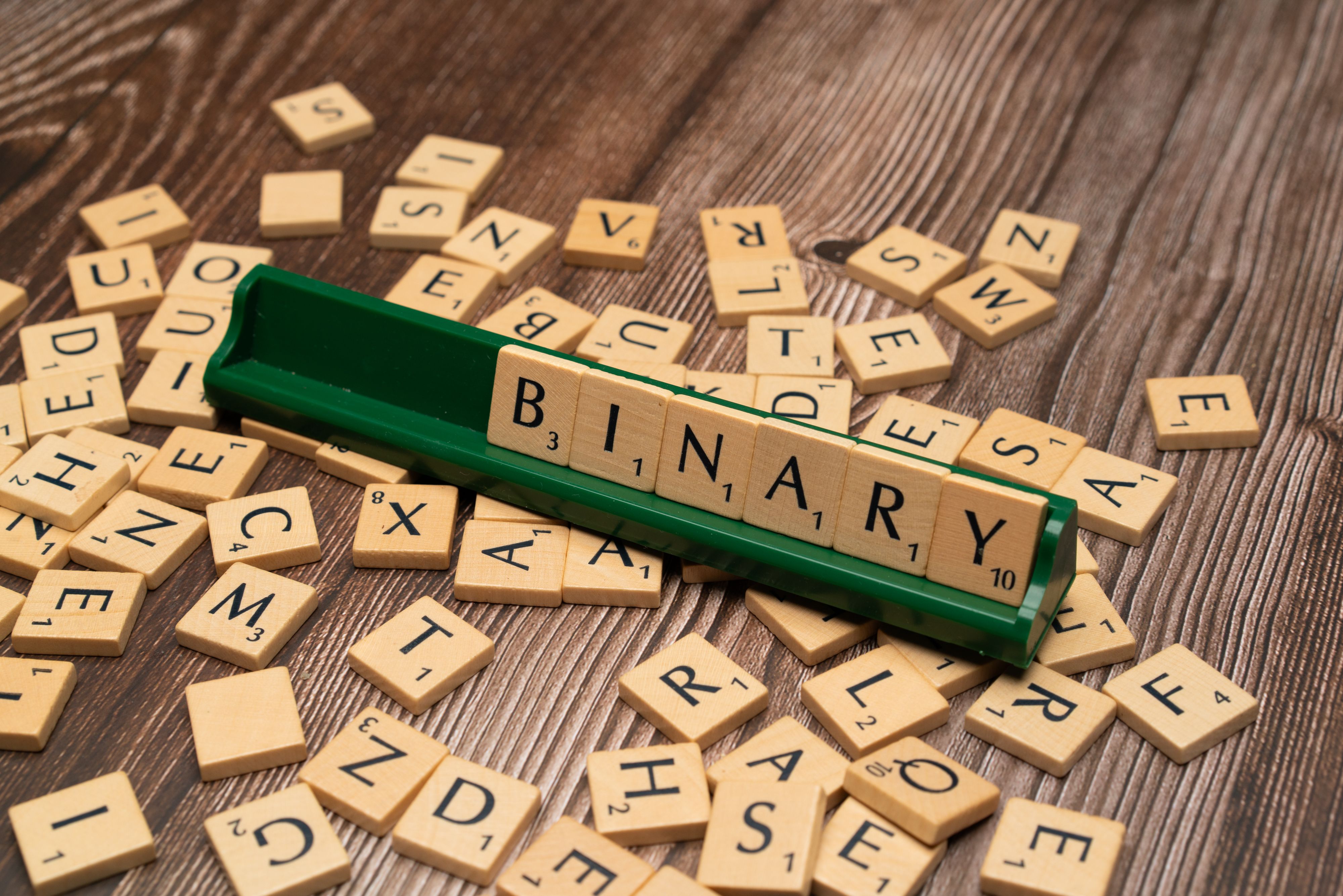 Close-up of scrabble tiles spelling 'Binary' surrounded by scattered letters on a wooden background.