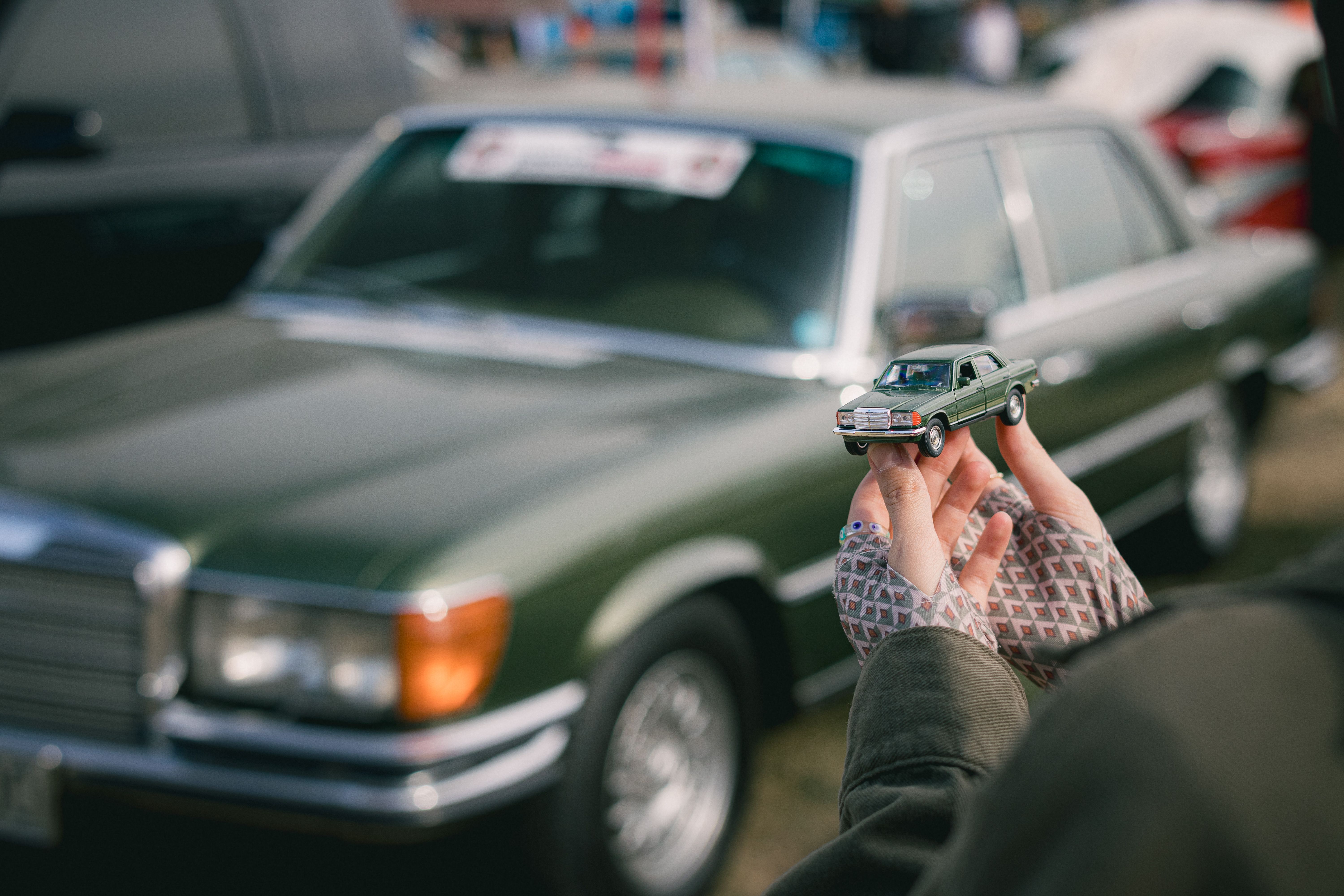 A green Mercedes-Benz W116 with a matching toy car held in hands outdoors.
