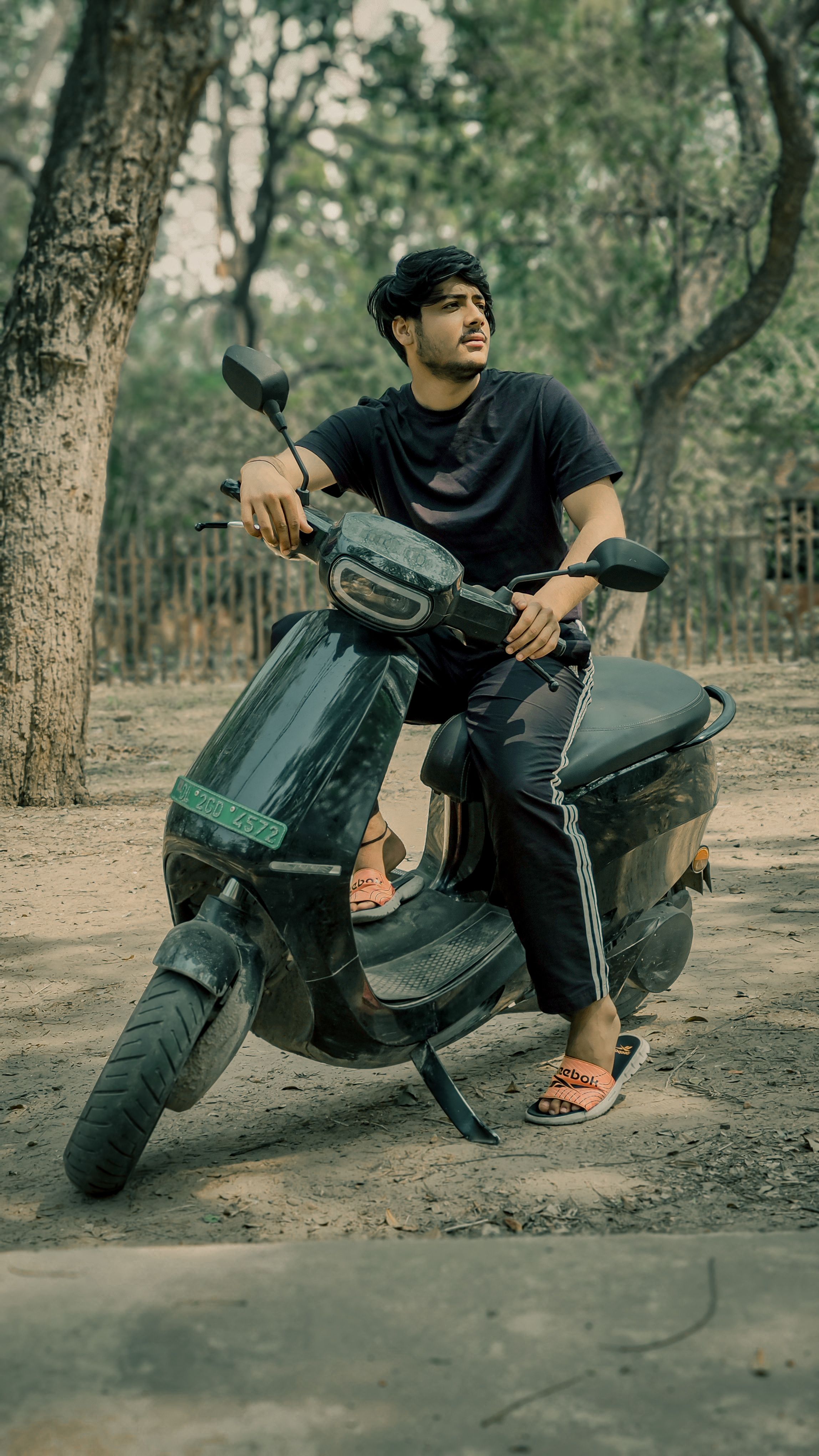 A young man in casual attire sits on a scooter in a park, conveying a relaxed lifestyle.