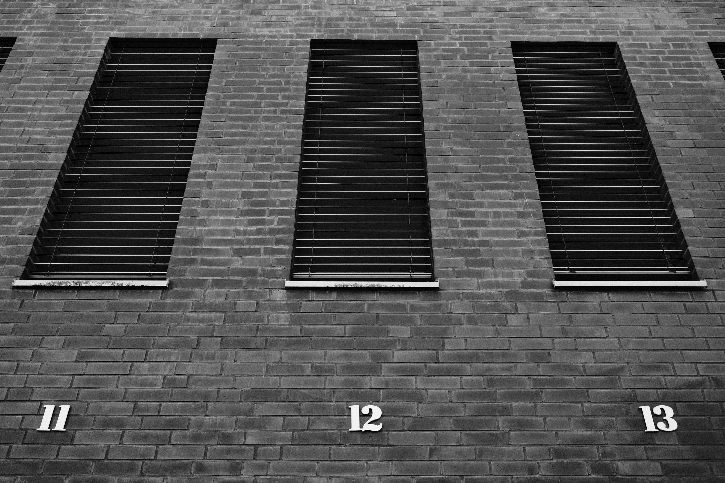 Monochrome image of a brick wall featuring three windows with closed blinds and numbers 11, 12, 13 below.
