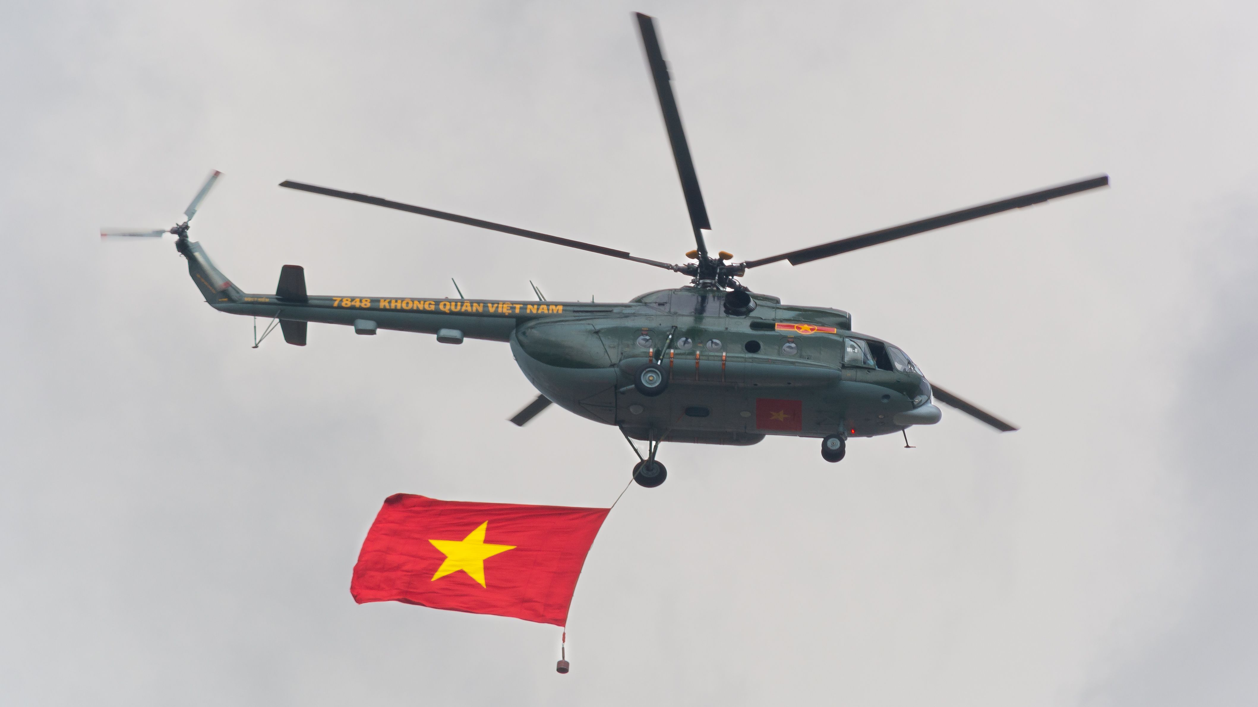 Vietnamese military helicopter flies with national flag during aerial display against gray sky.