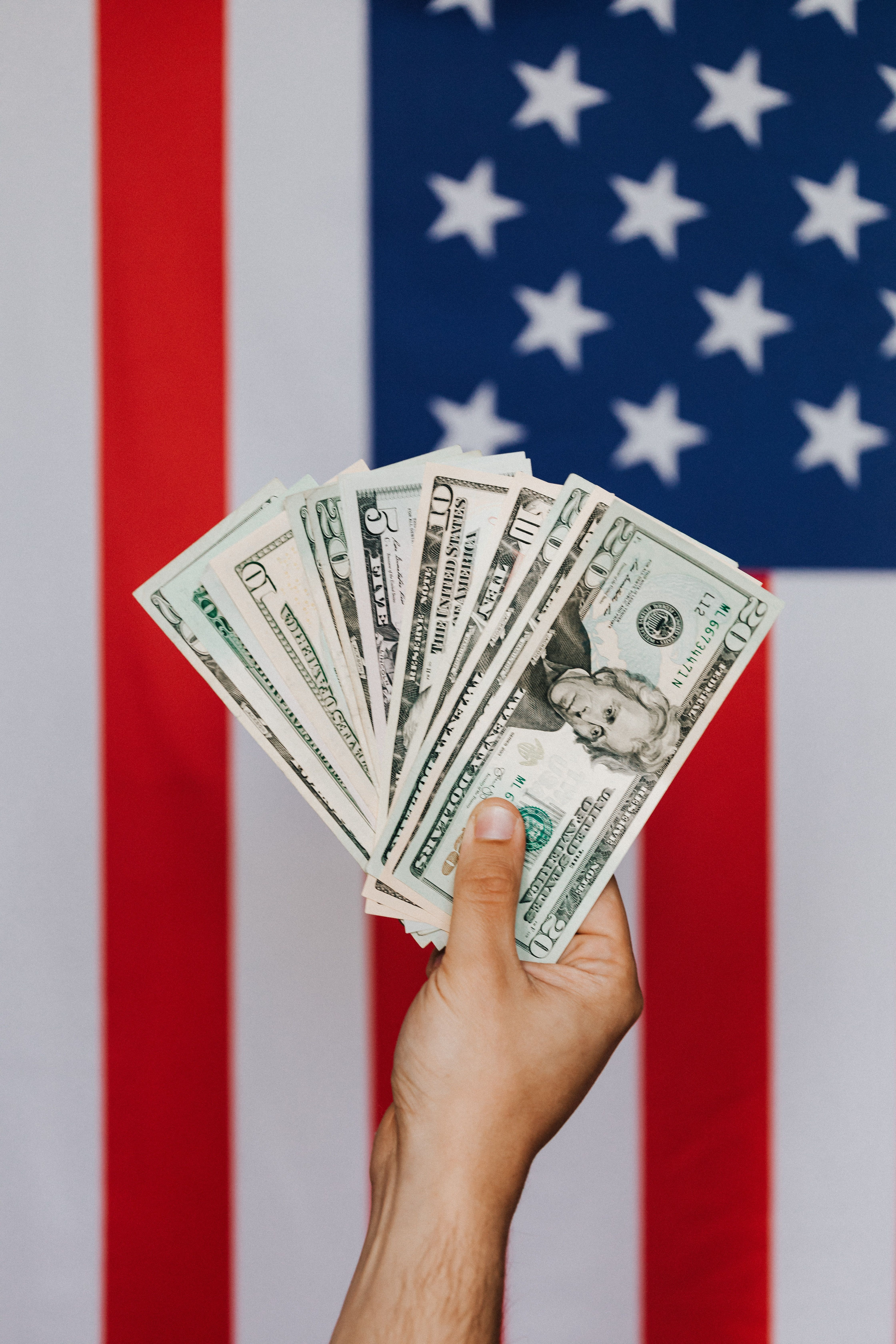 Crop unrecognizable person holding pile of dollars demonstrating money against national flag of United States of America