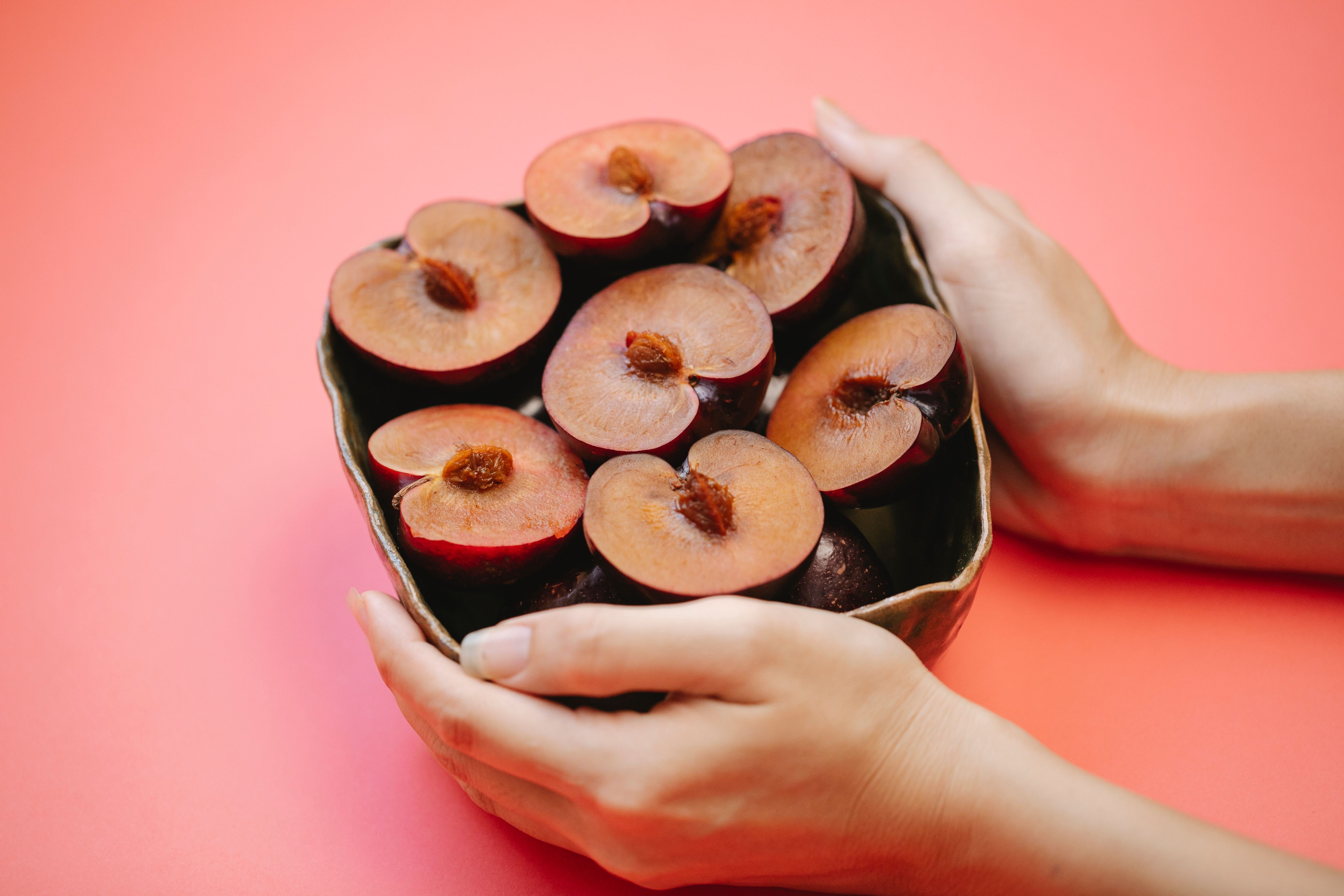 From above of crop faceless woman holding metal vase with ripe halved plums against pink background
