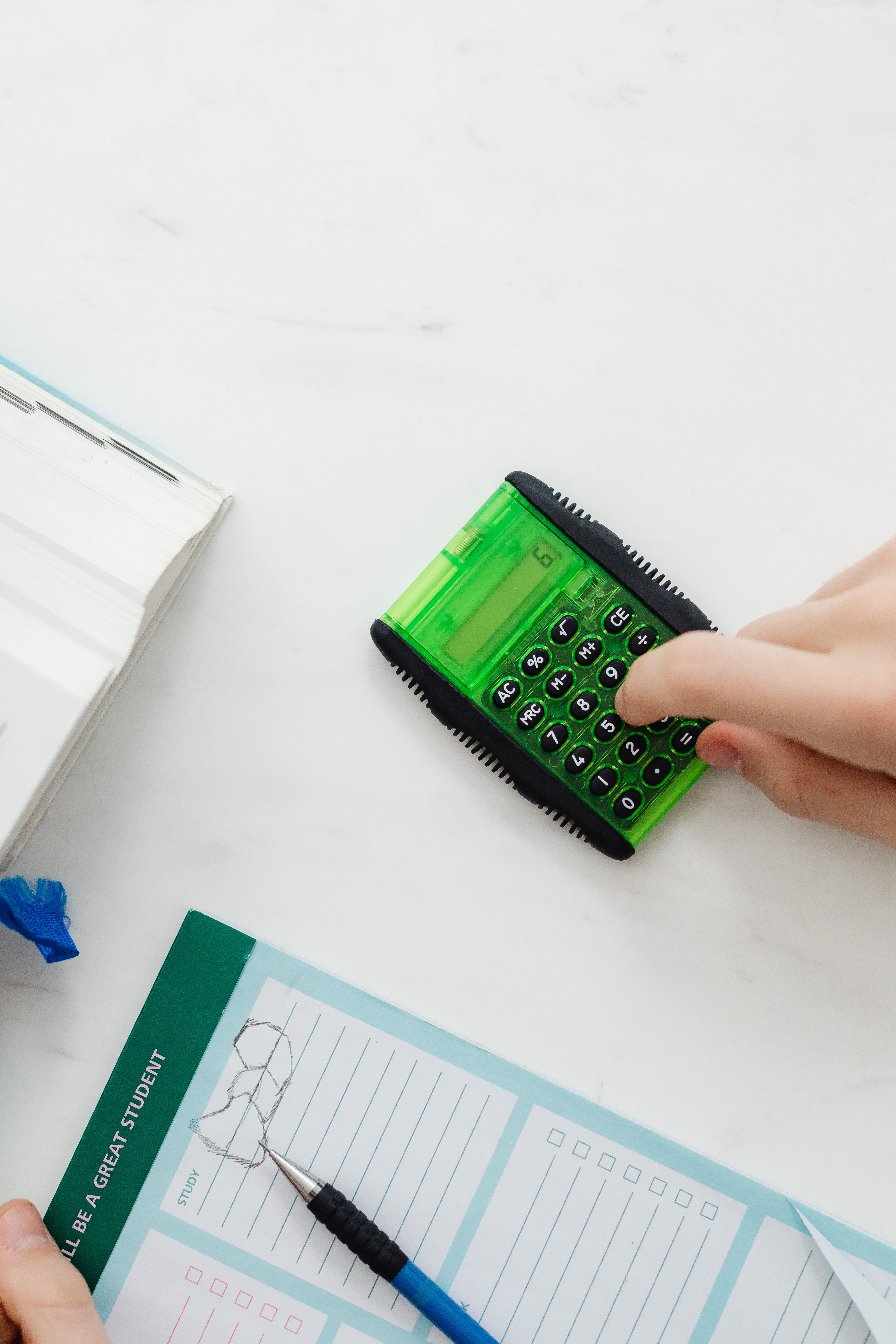 Close-up of hand using a green calculator on a study desk with a book and notebook.