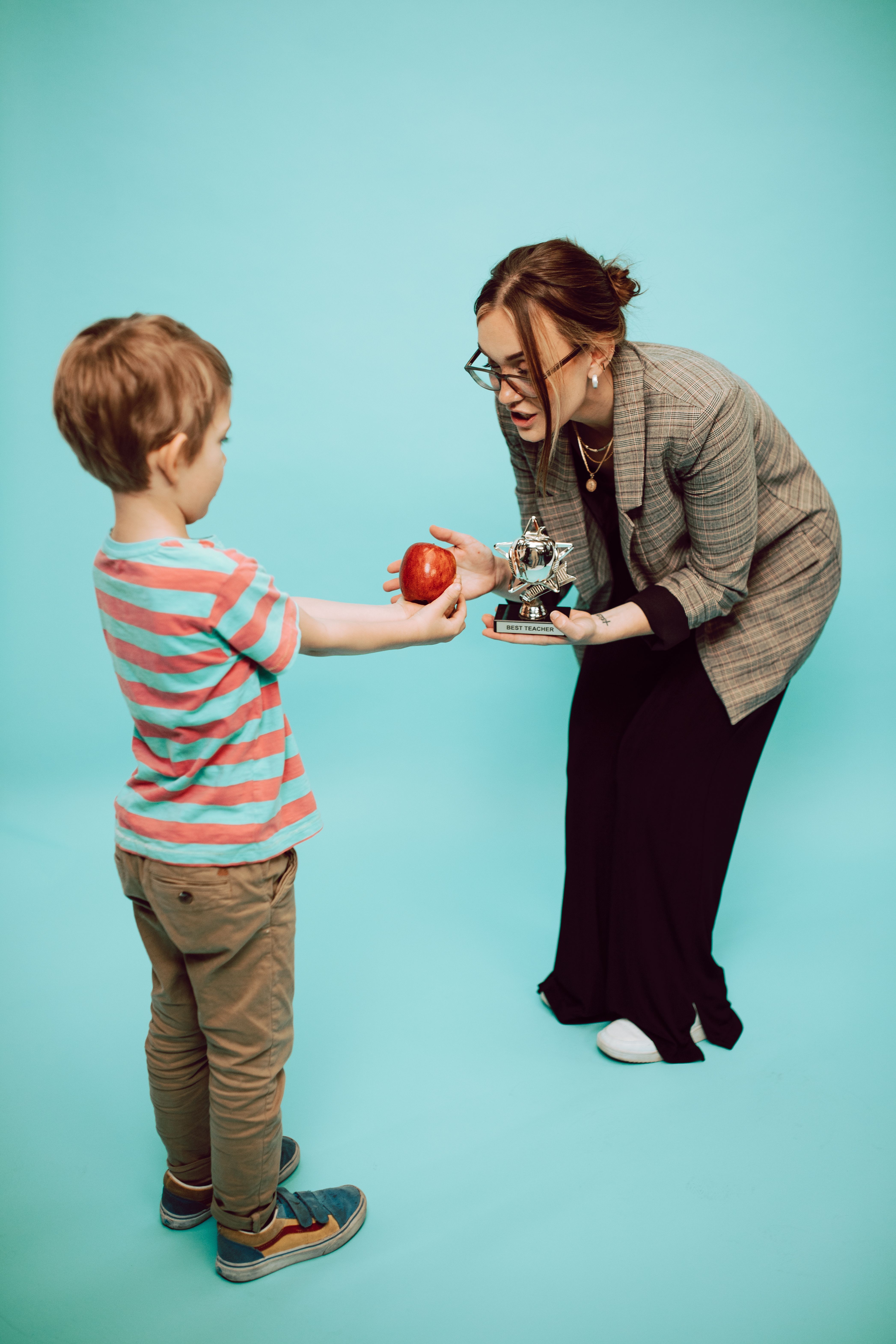 Young boy offers an apple to a smiling teacher holding a trophy indoors.
