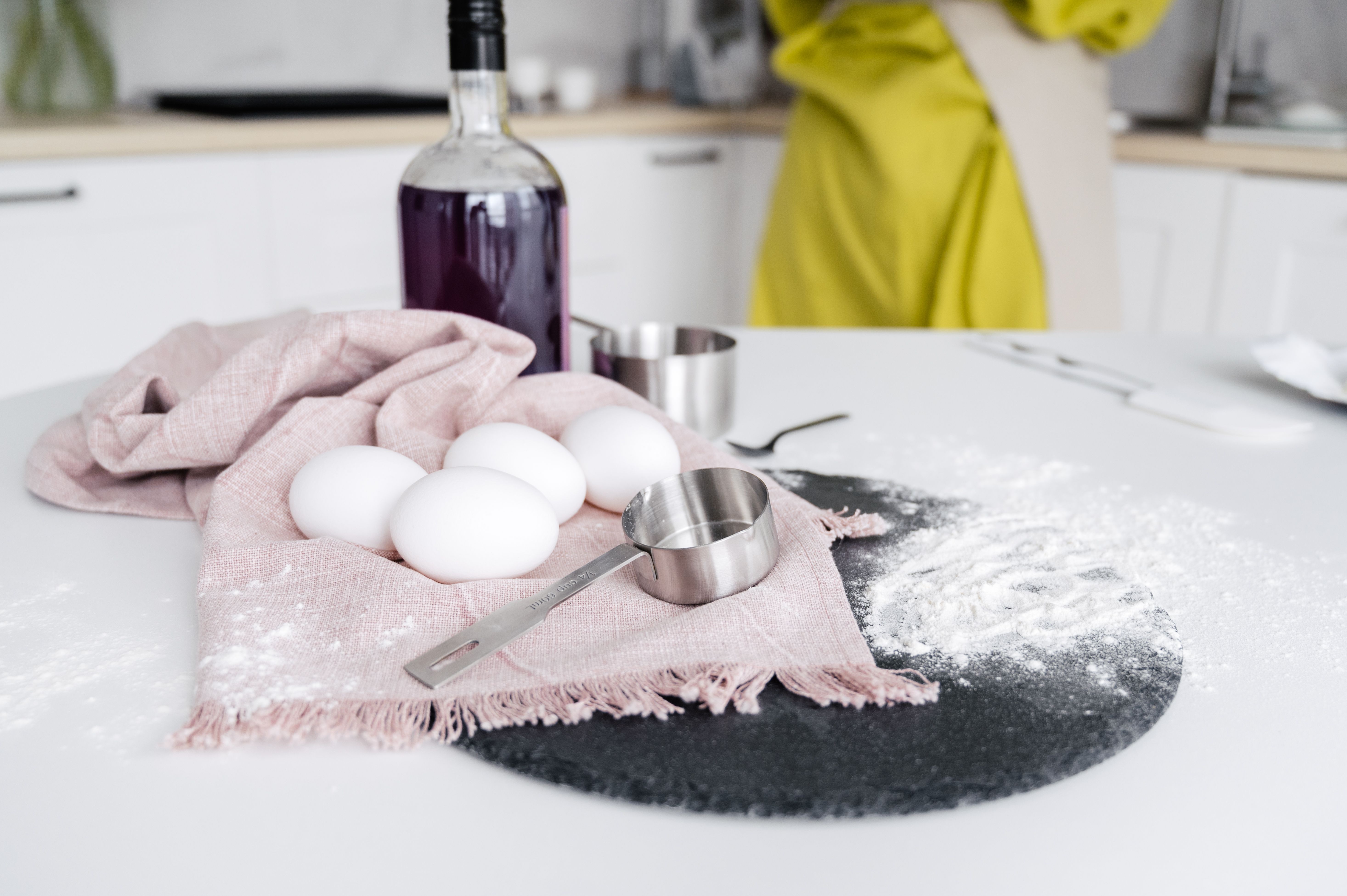 Crop unrecognizable female standing near white table with eggs and ladle placed on cloth with flour and glass bottle in modern kitchen